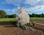 A small greenhouse with a white plastic cover and wooden frame stands outdoors in a grassy field under a blue sky with clouds. The greenhouse is surrounded by mulch, small potted plants, and a wooden bench in the foreground.