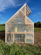 Small wooden greenhouse with transparent covering in a field under a blue sky.