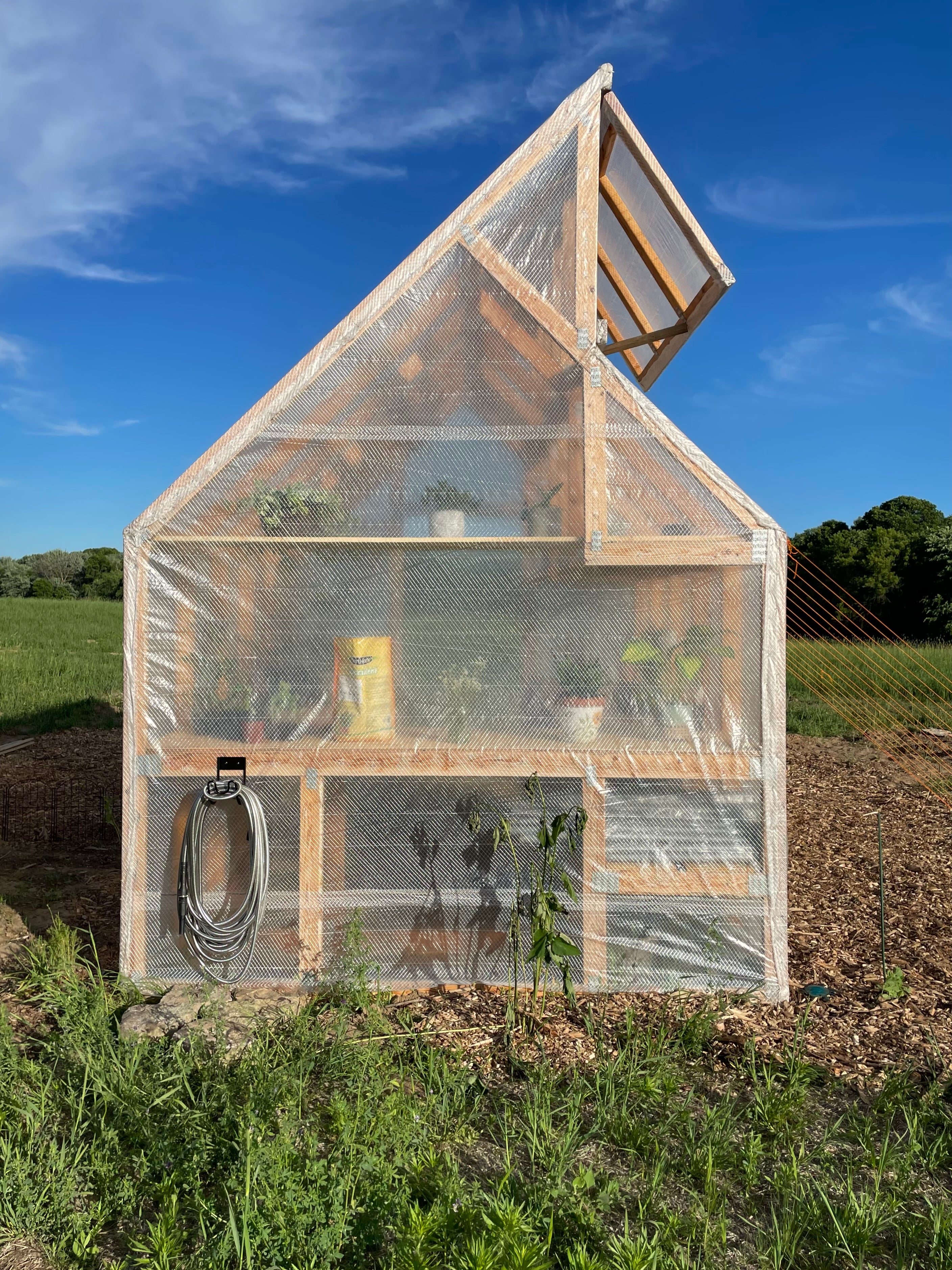Small wooden greenhouse with transparent covering in a field under a blue sky.